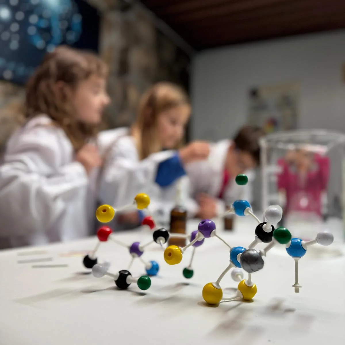 Modelos moleculares sobre una mesa con niños al fondo durante una actividad de ciencia.